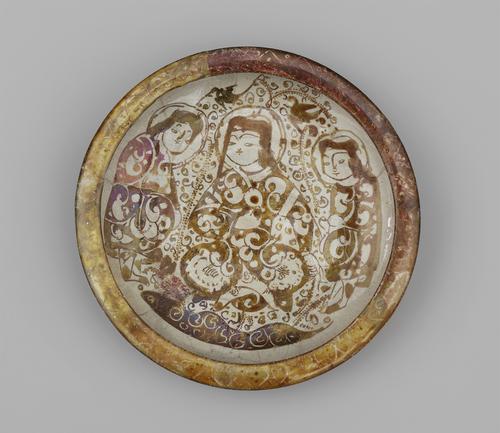 Shallow, decorated dish. The inside is decorated with three female figures in patterned robes, with swirling decoration around them. The exterior of the dish is inscribed with calligraphy, and the rim is decorated in yellow-brown with faint swirling marks.