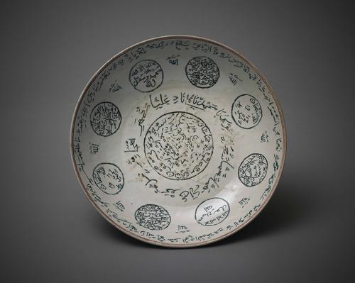 Top view of an off-white dish. In the centre is a green circle filled with writing, and a longer inscription wrapping around it. The sides are decorated with 8 similar green, text-filled circles, with small annotations between them. The rim is decorated with thin red lines and a long inscription.