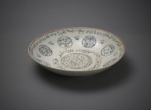 Side view of an off-white dish. In the centre is a green circle filled with writing, and a longer inscription wrapping around it. The sides are decorated with 8 similar green, text-filled circles, with small annotations between them. The rim is decorated with thin red lines and a long inscription.