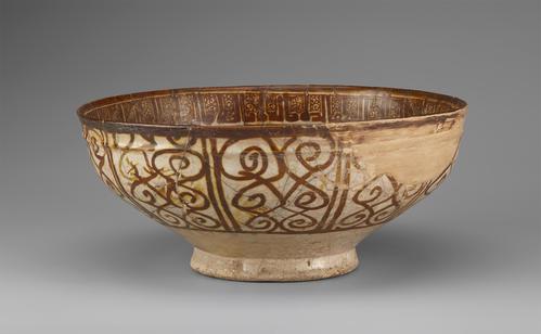 Side view of a circular bowl, decorated in brown paint over a cream background. The base is small and raised. The rim of the bowl is decorated in a band of brown script. The exterior is decorated with brown swirls.