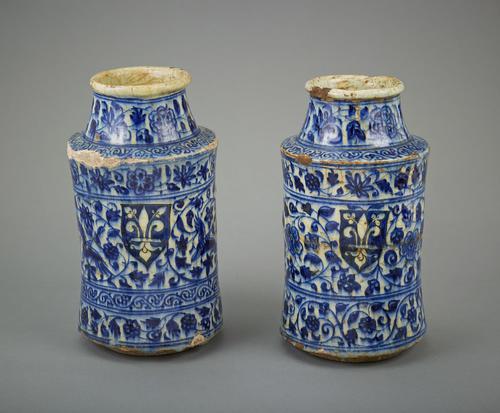 White ceramic jar with blue decoration, cylindrical form with sloping shoulder and short waisted neck with everted rim. The body has a shield cartouche enclosing the armorial device of the city of Florence reserved on a blue ground, and bands of floral scrolls.