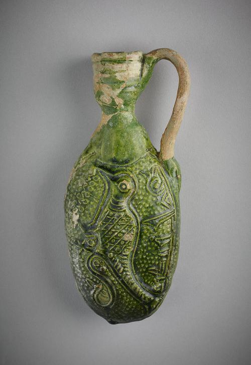 Flask, oval form, with handle attached on the body and rim of the mouth, made of greyish-white clay with dark green glaze. The body has a carved  design of of a bird under an inscription in kufic. Glaze flaked off handle and rim of mouth.