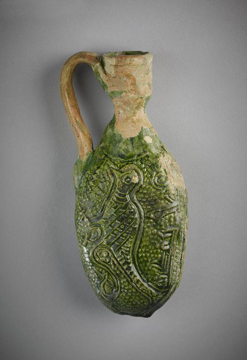 Back side of a flask, oval form, with handle attached on the body and rim of the mouth, made of greyish-white clay with dark green glaze. The body has a carved  design of of a bird under an inscription in kufic. Glaze flaked off neck, handle and rim.