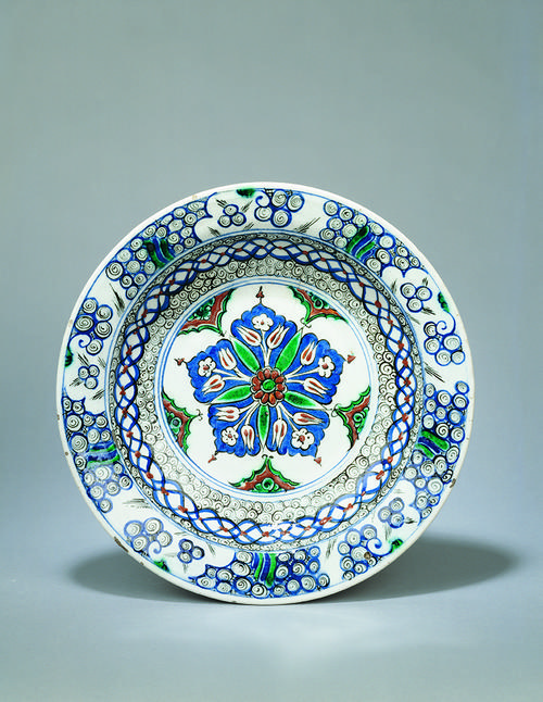 Dish of shallow rounded form, decorated in underglaze cobalt blue, green, relief red and outlined in black with a central design of radiating leaves, tulips and prunus blossom emanating from a floral rosette enclosed within a blue cartouche.