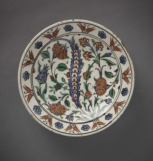 Dish, with a tall undulating lue flower in the centrewith red carnations, red rose buds and other blue an red flowers wave around it. A row of double red tulips and single blue flowers decorates the rim. 