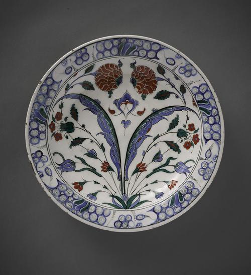 White plate decorated with two slender blue flowers out of a single plant, while behind it blue tulips, red carnations and other flowers, red and blue. Around the rim are blue whorls and the exterior of the dish is decorated with green and blue rosettes and trefoils.