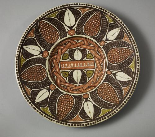 Dish, the centre is a small red band with a simple white Kufic inscription, a repetition of the name of God, “Allah”. Above and below it are identical leaf designs in dark brown and green. The central motif is circled by a red braid. 