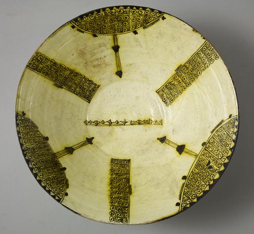 Bowl, reddish clay and white slip of the bowl have underglazed decorations drawn in dark brown and stained yellow. Wedge-like signs, form a single line in the centre. On the side, three large arcs separated by three vertical frames, all filled with peacock’s eyes.