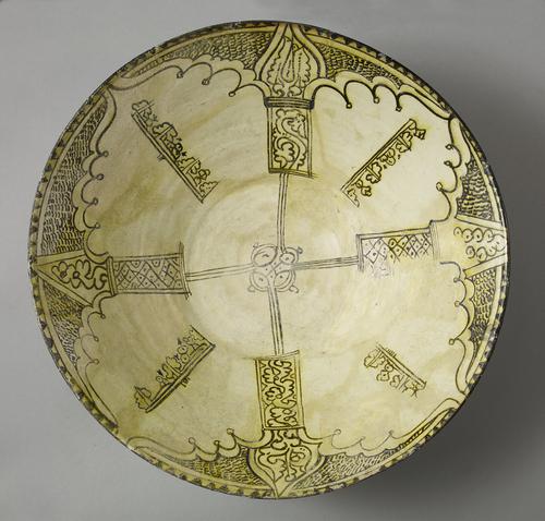 Dish, decorated with painting in dark brown, stained yellow, on a white slip shows. Four arc shapes at the rim, from which two onion-shapes in two of the arcs wide bands project filled with wavy lines. Each arc is separated from the next by a line of pseudo-Kufic.