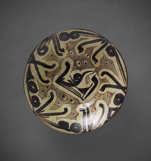 Bowl, with reddish brown clay and an ivory slip. Decorated on the sides with an undulating Kufic script in dark brown. The space between the letters is filled with angular areas of small dark brown dots and peacock’s eyes bounded by a red line.