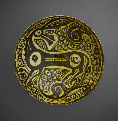 Bowl, decorated with dark brown, bright yellow and green. An deer like animal in dark brown and outlined in yellow, stands in the centre of the bowl and faces left. Around it yellow peacock’s eyes with green centres.