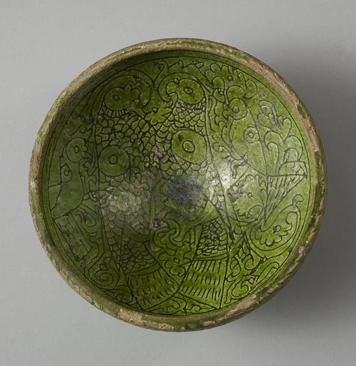 Bowl with green glaze and carved decoration of two large parrots facing each other,  to the left of the bowl’s centre. A smaller parrot stands over the back each of the larger birds. The background is filled with thick arabesques.