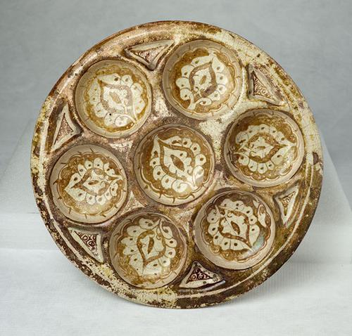 This large low dish contains seven hollows, six grouped around a central one. It is painted in brown lustre on a white ground. 