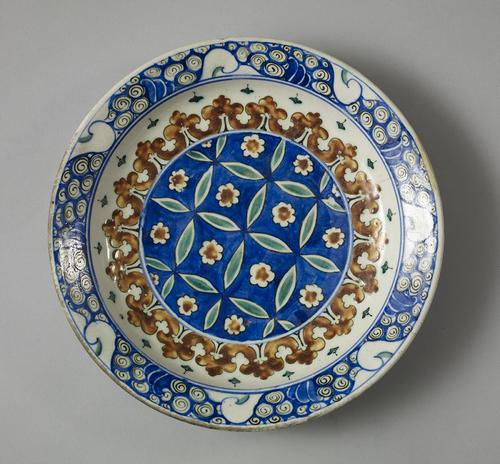 Dish, in the center is a medallion with a blue background which is crossed by a pattern of light green ovals and red and white flowers. A red scrollwork pattern encloses the center design while the rim of the dish is covered with the wave and whorl pattern.