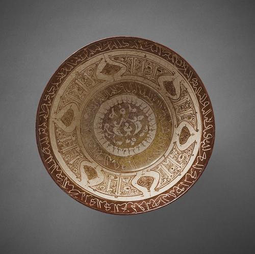 Bowl decorated in brown lustre on a white ground. A brown circle with white arabesques is in the centre. Around it are a bands of white inscription, a wider band of Kufic letters, leaves and vine scrolls and a white Naskhi inscription on a brown background decorates the rim. 