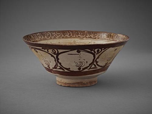 A bowl decorated in brown lustre on a white ground. A white Naskhi inscription on a brown background decorates the rim. Seven large, rough, brown circles decorate the underside.