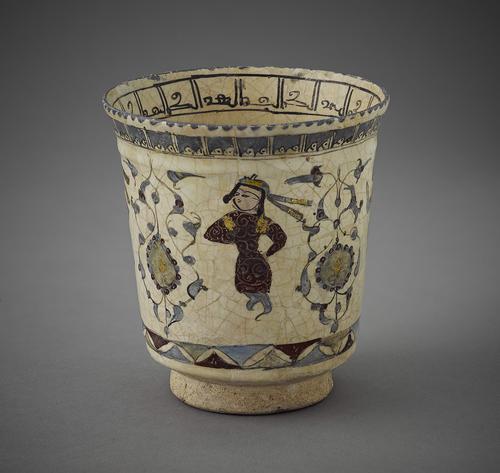 Beaker with a man dressed in a long red garment on a white background on either side of him foliage. The lower part of the sides decorated with a band of purple and blue triangles, the top with a band of white. The inside rim is ornamented with a band of black Kufic script.