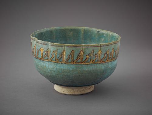 Bowl with a low, unglazed foot and vertical, fluted sides has an opaque, turquoise glaze and is decorated along the outside rim with a row of repeating Kufic letters in gold, outlined in red.