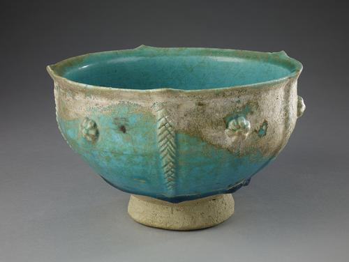 Bowl with flaring rim, turquoise glazed, silvery iridescence. The foot is unglazed.  On the outside, five vertical, appliqué strips of clay, not equidistant from each other, are cross-hatched like strands of rope and end in points projecting out from the rim and single, small rosett between them.