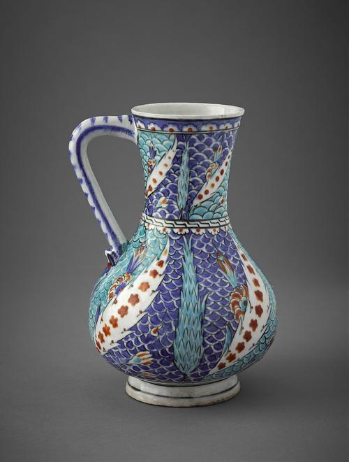 Pottery jug, the body is pear-shaped and the neck flares slightly. Around the foot is a single, blue line. The body and the neck are covered with light and dark blue scales as a background against which stand blue cypress trees, various flowers, and tilting white trees with red blossoms.