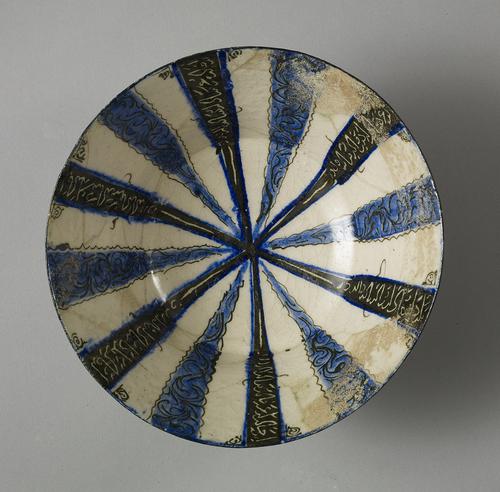 White bowl decorated in blue and black. Twelve stripes radiate from the centre of the bowl: six are blue with black borders and black waterweed decoration, the other six are black with blue borders and white inscriptions.