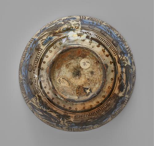 Bottom of a bowl, the exterior sides contain drawings in brown on a cream ground with some light blue colouring for water and some elements of the landscape. On the foot of the bowl are two round stickers.