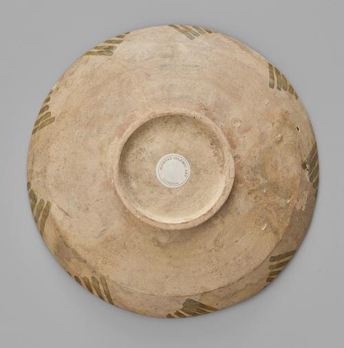 Bottom of a shallow bowl, view of the round foot with a white round sticker in the center, and outside walls decorated with repetative green lines in a triangular design.